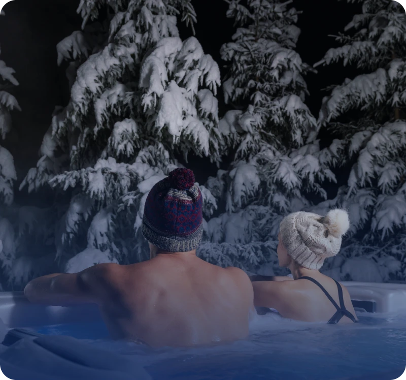 Two people relaxing in a hot tub surrounded by snowy trees at night.